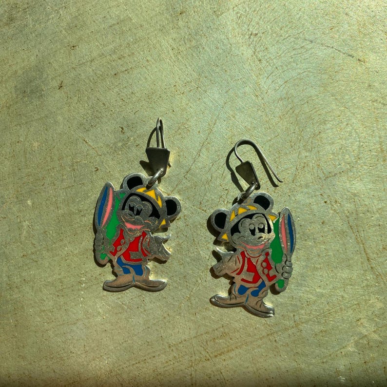 Op de afbeelding: Een paar zilverkleurige oorbellen met een cartoon muis karakter. De muis draagt een gele hoed, een rode jas, een blauwe broek en houdt een kleurrijk surfboard vast. De oorbellen hebben een haaksluiting.