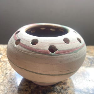 May include: A round, off-white ceramic vessel featuring a wide opening at the top surrounded by a series of small, evenly spaced circular holes. The exterior surface displays subtle, swirling horizontal bands of muted pink and sage green clay.