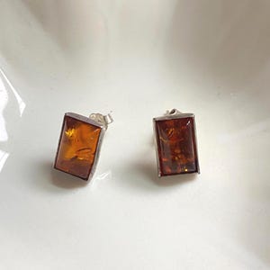 May include: A pair of rectangular amber stud earrings with a silver-colored border. The amber has a warm, translucent, honey-brown color. The earrings are displayed on a white surface.