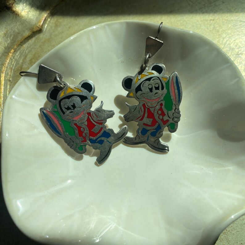 Op de afbeelding: Een paar vintage Mickey Mouse-oorbellen. Elke oorbel heeft een zilverkleurige Mickey Mouse-figuur in een kleurrijke outfit, met een veer. De oorbellen hebben een oorhaaksluiting.