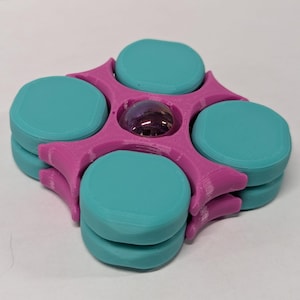 May include: A 3D-printed fidget spinner in a vibrant color scheme. The spinner features a magenta frame with five teal-colored circular pads. A small, iridescent ball sits in the center. The design is symmetrical and modern.