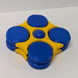 Puede incluir: Un juguete fidget impreso en 3D con un diseño vibrante. Tiene una estructura central amarilla con cuatro componentes circulares azules apilados. Una pequeña esfera azul se encuentra en el centro.