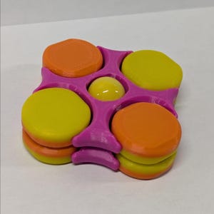 May include: A colorful puzzle toy featuring a magenta frame with circular cutouts. The toy has orange and yellow discs stacked in the cutouts, and a yellow sphere in the center. The discs are stacked in pairs.