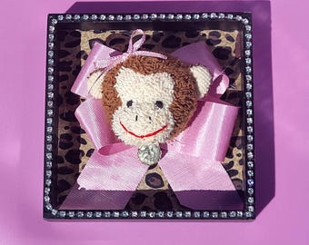 Op de afbeelding: Een vierkante zwarte lijst met een strass-rand bevat een bruin en crème pluche apengezicht gecentreerd op een roze lintstrik. De achtergrond in de lijst heeft een bruin en zwart luipaardprintpatroon.
