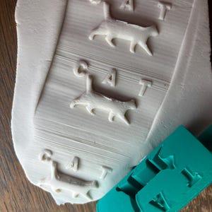 May include: A white, flat, rectangular piece of clay featuring three embossed impressions of a cat silhouette and the word CAT, accompanied by a teal plastic rolling pin tool with matching recessed cat and letter shapes for imprinting.