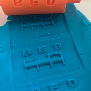 May include: An orange plastic rolling pin with a raised pattern of the word BED and a bed icon, shown pressing into a flat sheet of blue modeling clay to create a repeating embossed impression of the text and the bed graphic across the surface.