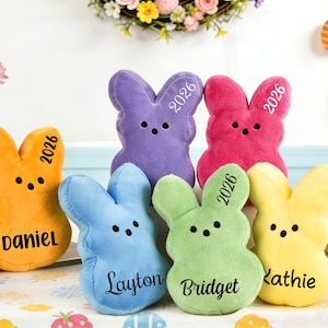 Puede incluir: Seis conejos de peluche de colores: naranja, morado, rosa, azul, verde y amarillo. Cada conejo tiene un nombre y el año 2026 impreso. Los conejos están colocados sobre una mesa con decoraciones de Pascua.