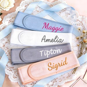 May include: Four personalized spa headbands in blue, white, gray, and peach, each embroidered with a different name in a complementary color. The headbands are arranged on a lace doily with a blue ribbon and flowers.