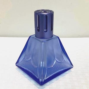 Peut inclure: Lampe à parfum en verre bleu avec une base géométrique en forme de pyramide et un dessus cylindrique perforé. La lampe a un aspect givré et est conçue pour diffuser des parfums. Le design général est moderne et décoratif.