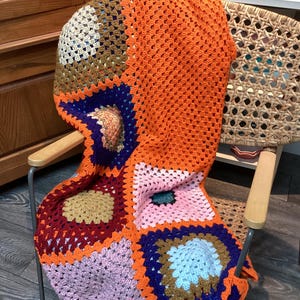 Peut inclure: Une couverture crochetée à la main, aux couleurs vives, drapée sur une chaise. Le plaid présente un motif patchwork avec des carrés orange, bleu, rose et marron. Un article décoratif et confortable.