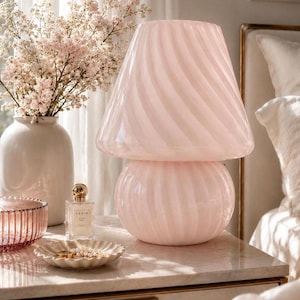 Peut inclure: Une lampe de table en forme de champignon fabriquée en verre rose pâle avec un motif côtelé en spirale subtil. La lampe présente un large abat-jour évasé reposant sur une base arrondie et bulbeuse, créant un design monochrome aux tons pastel.