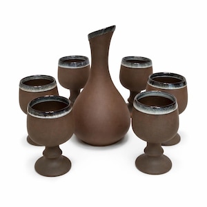 Peut inclure: Ensemble de service à boisson en céramique marron comprenant une carafe élancée et six gobelets assortis. Les gobelets ont une forme arrondie avec une tige et une base. Les bords des gobelets et le bec de la carafe ont une bordure sombre contrastante.