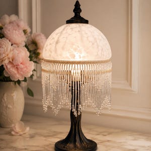 Peut inclure: Une lampe de table avec une base couleur bronze foncé et un abat-jour en verre blanc moucheté en forme de dôme. L'abat-jour est orné d'une bordure en tissu beige et de longues franges de perles en verre transparent en festons.