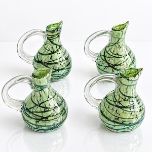 Peut inclure: Quatre petits pichets décoratifs en verre avec des anses et des becs clairs. Les pichets sont de couleur vert clair avec des lignes tourbillonnantes vert foncé et noires. Les pichets sont exposés sur une surface blanche.