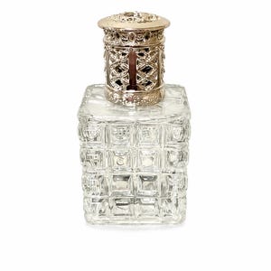 May include: A clear glass fragrance diffuser bottle with a square base and a grid-like textured pattern. The bottle is topped with a metallic silver-toned decorative cap featuring an intricate openwork design with small cutouts.