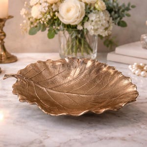 Peut inclure: Un plateau décoratif doré en forme de feuille. Le plateau présente des nervures détaillées et une surface texturée. L'arrière-plan comprend des bougies dans des bougeoirs dorés, un vase de fleurs, des livres et un flacon de parfum. Un accessoire décoratif.