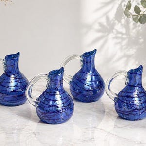 Peut inclure: Quatre carafes en verre bleu cobalt avec des anses en verre transparent. Chaque carafe a un corps arrondi avec des lignes tourbillonnantes bleu foncé. Les carafes sont disposées sur une surface blanche, avec un vase de fleurs séchées et de la verdure en arrière-plan.