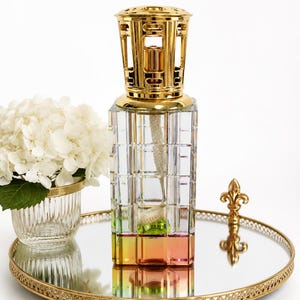 May include: A square glass fragrance lamp with a faceted grid pattern and an iridescent base, topped with a polished gold-tone metal crown. The lamp sits on a round mirrored tray with a gold-tone filigree edge, next to a small glass vase.