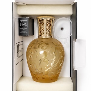 Peut inclure: Lampe à parfum décorative ambrée avec un dessus en filigrane doré, présentée dans une boîte avec un entonnoir et une petite boîte noire étiquetée "Elina". La lampe a un motif floral gravé.