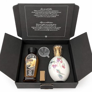Peut inclure: Un coffret cadeau noir contenant une lampe parfumée blanche et dorée avec un motif floral, une bouteille d'huile parfumée, un entonnoir et un petit objet doré. La boîte contient du texte en français et en anglais.