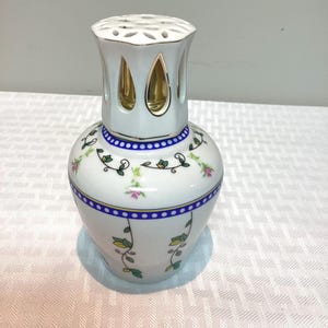 Peut inclure: Lampe à parfum en céramique blanche avec un motif floral. La lampe a un corps bulbeux avec une bordure bleue et dorée, et un dessus perforé. Le motif comprend des fleurs roses et des feuilles vertes. La partie supérieure a des accents dorés.