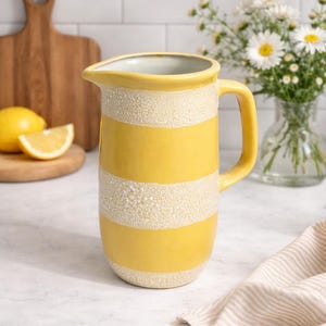 Peut inclure: Pichet en céramique à rayures jaunes et blanches avec une poignée. Le pichet est posé sur un comptoir blanc avec une planche à découper en bois, des citrons et un vase de fleurs blanches en arrière-plan.