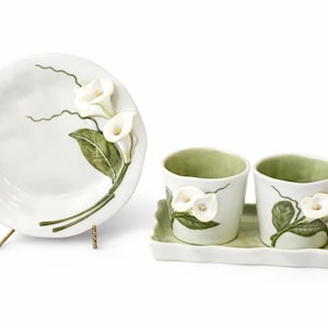 Peut inclure: Une assiette de service en céramique blanche et deux petites tasses avec un plateau assorti. Chaque pièce est décorée de callas blanches et de feuilles vertes. L'assiette est sur un support en métal doré.