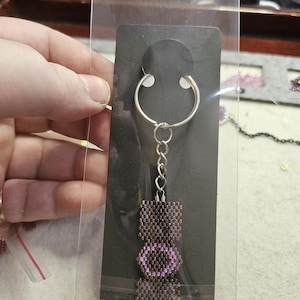May include: A beaded keychain featuring a rectangular design with dark brown beads and a central circular pattern of light purple beads. The keychain is attached to a silver metal split ring and chain, presented on a black display card in a clear sleeve.