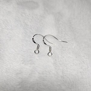 以下が含まれることがあります： 銀色のイヤリングフックのペア。フックは湾曲しており、下部に小さなループがあり、ループのすぐ上にコイル状のデザインがあります。イヤリングは柔らかい白い背景に表示されています。