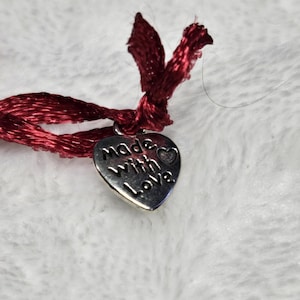 以下が含まれることがあります： 「made with love」の文字が刻まれた、小さな銀色のハート型のチャームのクローズアップ。チャームは、上部で結ばれた濃い赤色のサテンリボンに取り付けられています。