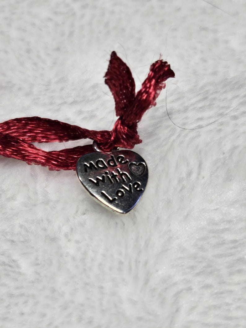 以下が含まれることがあります： 「made with love」の文字が刻まれた、小さな銀色のハート型のチャームのクローズアップ。チャームは、上部で結ばれた濃い赤色のサテンリボンに取り付けられています。