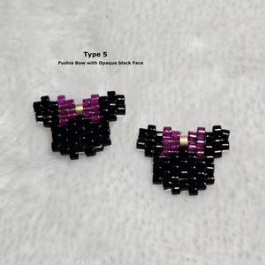 以下が含まれることがあります： 黒いビーズで作られた2つのリボン型アクセサリー。フクシアとゴールドのディテールが施されています。テキストには「Type 5」と「Fushia Bow with Opaque black Face」と書かれています。