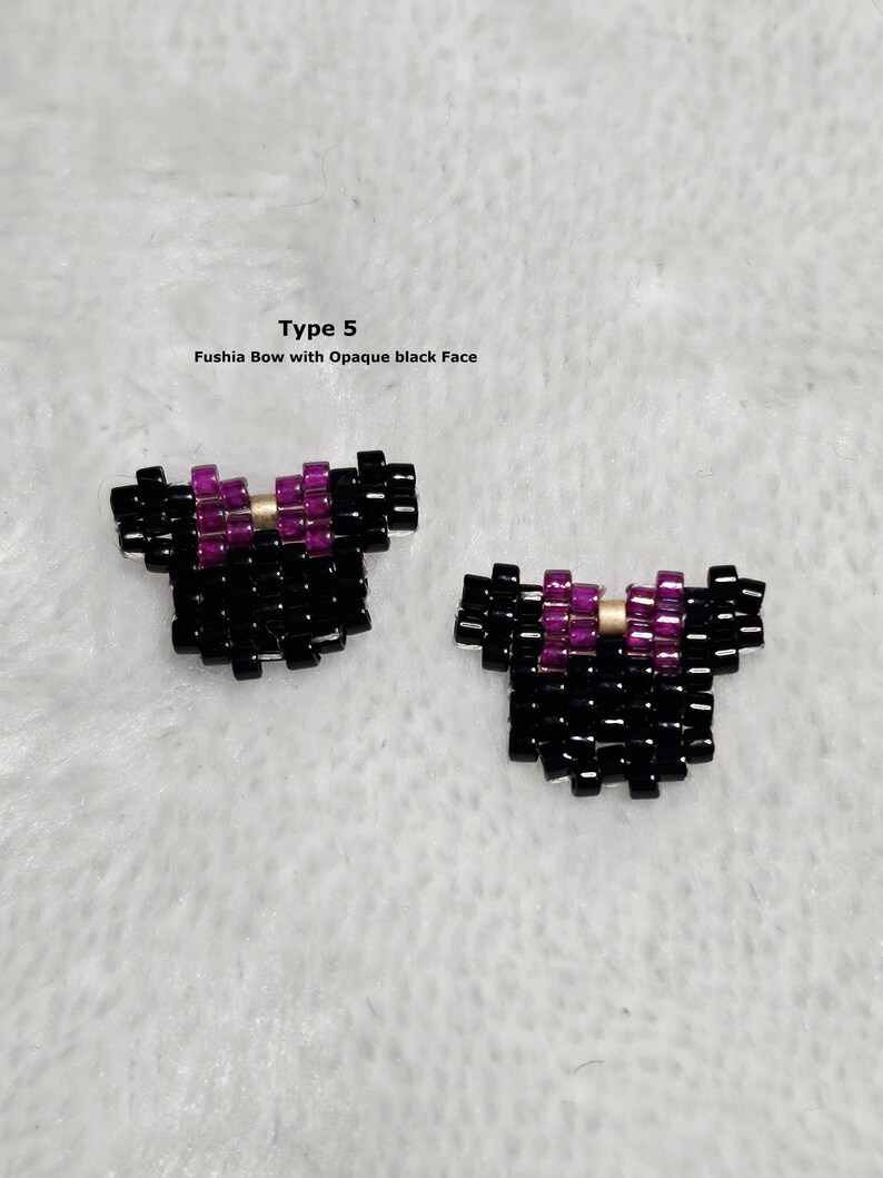 以下が含まれることがあります： 黒いビーズで作られた2つのリボン型アクセサリー。フクシアとゴールドのディテールが施されています。テキストには「Type 5」と「Fushia Bow with Opaque black Face」と書かれています。