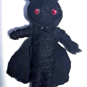 Puede incluir: Muñeca negra hecha a mano con ojos rojos, antenas y una pequeña capa. La muñeca está hecha de un material difuso y tiene una cadena adjunta en la parte superior para colgar. El diseño de la muñeca se asemeja a una polilla o insecto.