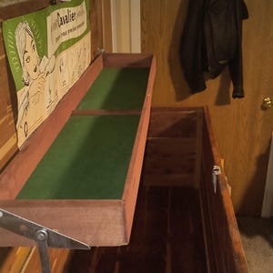 May include: A vintage wooden chest with a pull-out tray lined with green felt. A poster with a woman and the word "Cavalier" is visible. A black jacket hangs on a wooden door in the background. The chest is open, revealing its interior.