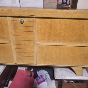 May include: A light brown wooden dresser with multiple drawers and a cabinet. A red and black tape measure sits on top. The open drawer reveals a red folder, cleaning supplies, and other items.