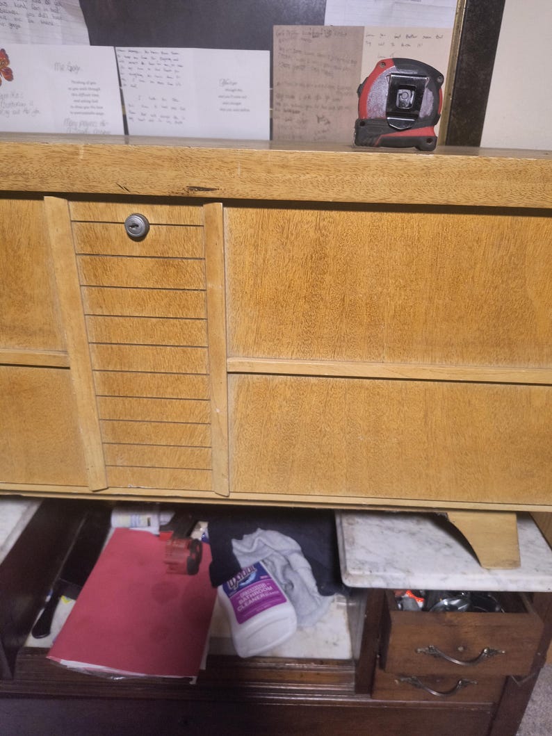 May include: A light brown wooden dresser with multiple drawers and a cabinet. A red and black tape measure sits on top. The open drawer reveals a red folder, cleaning supplies, and other items.