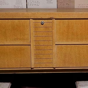 May include: A light brown wooden chest with a lock and keyhole. The chest has a rectangular shape with two drawers on each side and a central panel with vertical grooves. It stands on angled legs.