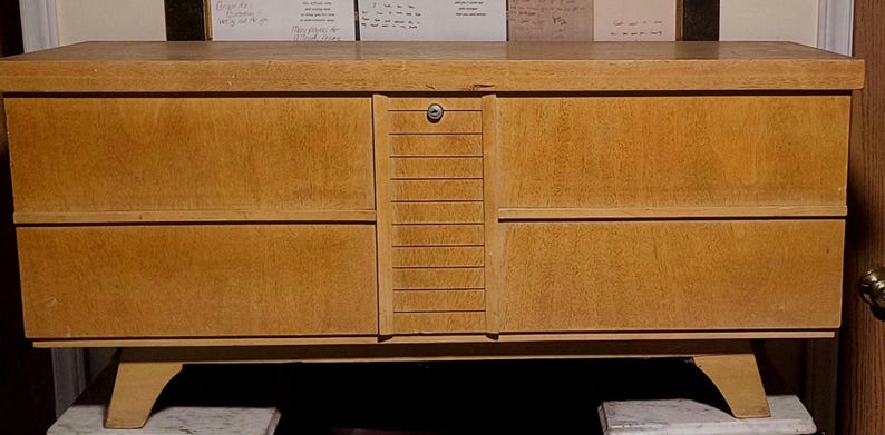 May include: A light brown wooden chest with a lock and keyhole. The chest has a rectangular shape with two drawers on each side and a central panel with vertical grooves. It stands on angled legs.