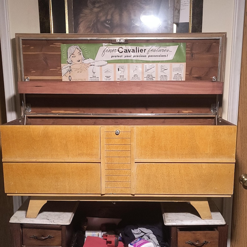 May include: A vintage wooden chest with a light brown finish, featuring a rectangular design with a central lock and two drawers. The chest has a hinged lid and is supported by angled legs. The interior includes a sign that reads "finer Cavalier features... protect your precious possessions!"