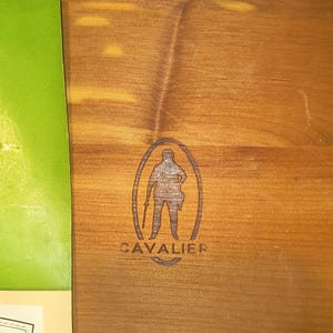 May include: A close-up of a wooden surface with a dark brown oval emblem. The emblem features a silhouette of a person holding a staff, with the word "CAVALIER" below. The wood has a natural grain.
