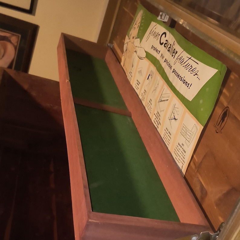 May include: Open wooden drawer with a green felt lining. A vintage advertisement is attached to the inside of the chest, with the text "finer Cavalier features... protect your precious possessions!"