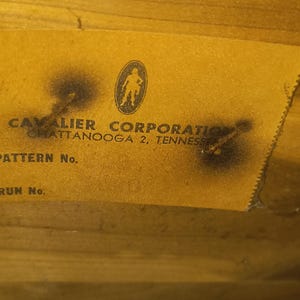 May include: Close-up of a yellowed label with the text "CAVALIER CORPORATION CHATTANOOGA 2, TENNESSEE" and a logo of a silhouetted figure. The label is affixed to a wooden surface, with visible wear and tear.