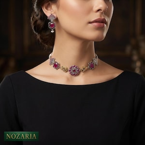 May include: A silver-toned necklace and matching earrings set with ruby-colored gemstones. The necklace features a floral design with gold accents. The earrings have a similar design, with a dangling pearl. The jewelry is worn against a black top.