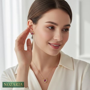 May include: A gold necklace and matching stud earrings set, each with a central red gemstone and surrounding clear stones. The pendant and earrings have a floral design. The brand name "NOZARIA" is visible in the bottom left corner. The jewellery is worn.