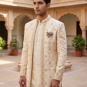 May include: A cream-colored sherwani featuring intricate tonal embroidery, a high mandarin collar, and a button-down front. An ornate elephant-shaped brooch with colorful accents is pinned to the chest. The garment is long-sleeved and structured.