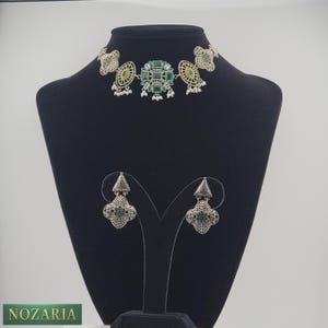 May include: A silver-toned necklace and matching earrings set. The necklace features a central design of green gemstones and a choker style. The earrings have a similar design. The jewelry is displayed on a black mannequin.