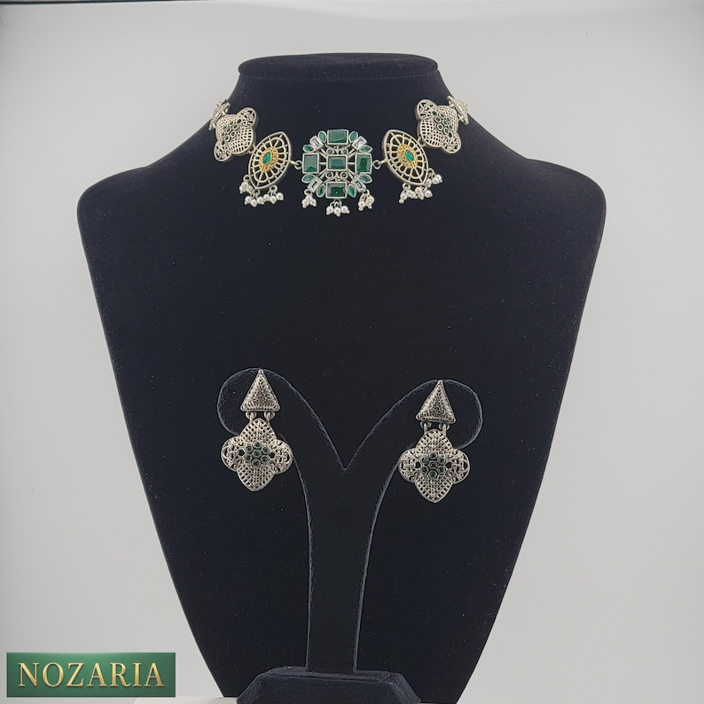 May include: A silver-toned necklace and matching earrings set. The necklace features a central design of green gemstones and a choker style. The earrings have a similar design. The jewelry is displayed on a black mannequin.