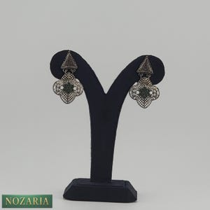 May include: Silver-toned filigree earrings with a geometric design. Each earring features a triangular top and a four-leaf clover-shaped bottom with green accents. The earrings are displayed on a black jewelry stand against a white background.