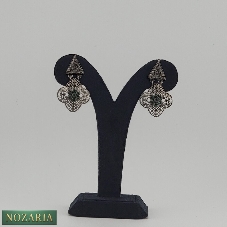 May include: Silver-toned filigree earrings with a geometric design. Each earring features a triangular top and a four-leaf clover-shaped bottom with green accents. The earrings are displayed on a black jewelry stand against a white background.